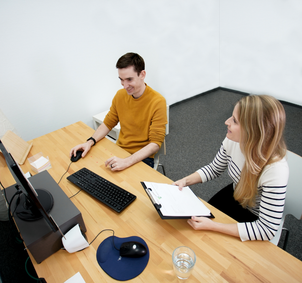 Zwei Mitarbeiter von Custom Interactions sitzen an einem Arbeitsplatz und lachen. Ein Mitarbeiter bedient lächelnd den Computer, während eine Mitarbeiterin lächelt und ein Klemmbrett in der Hand hält.
